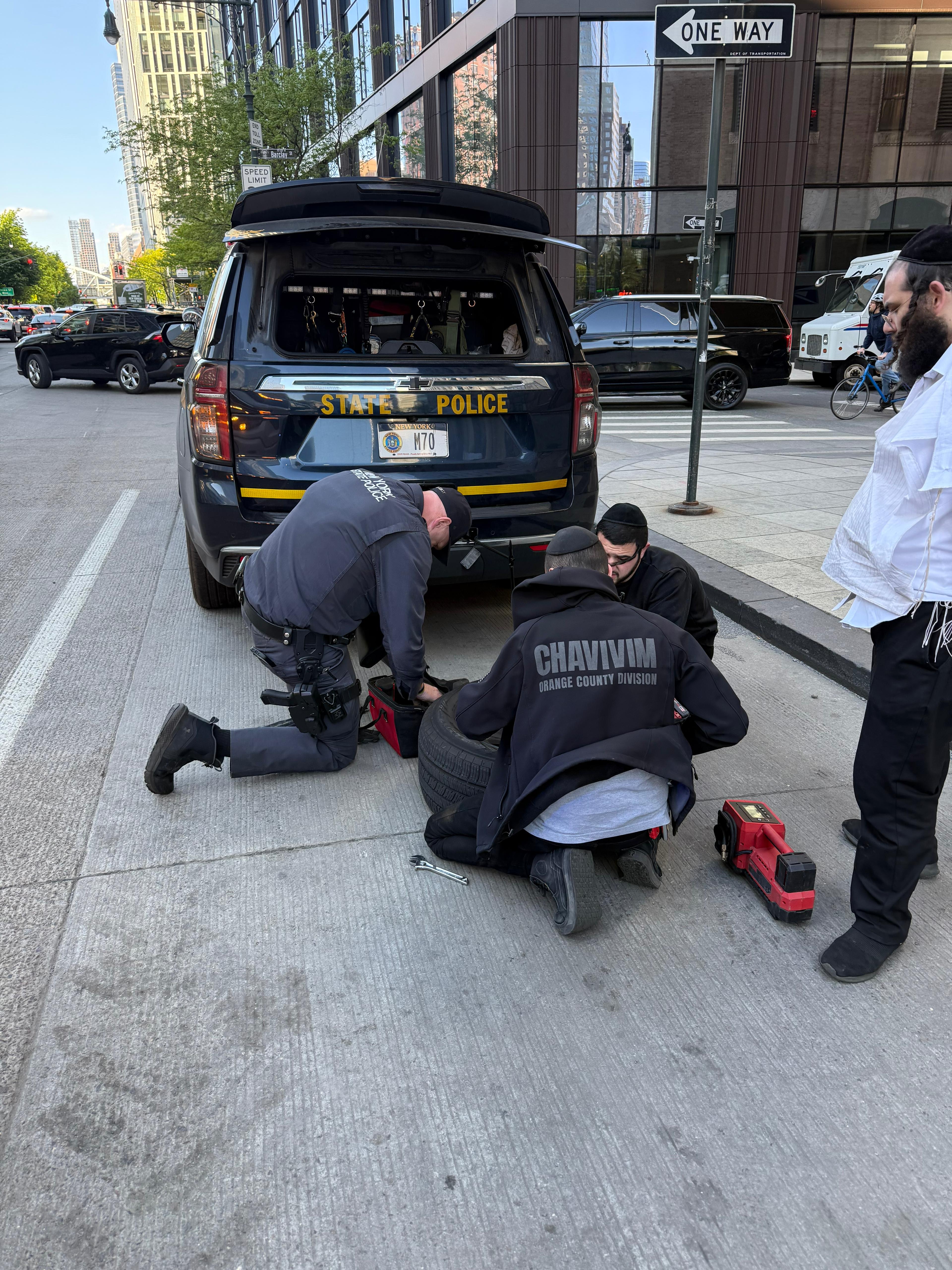 NYSP trooper and two Chavivim members crouched together at the rear wheel of the state police SUV on a Manhattan street