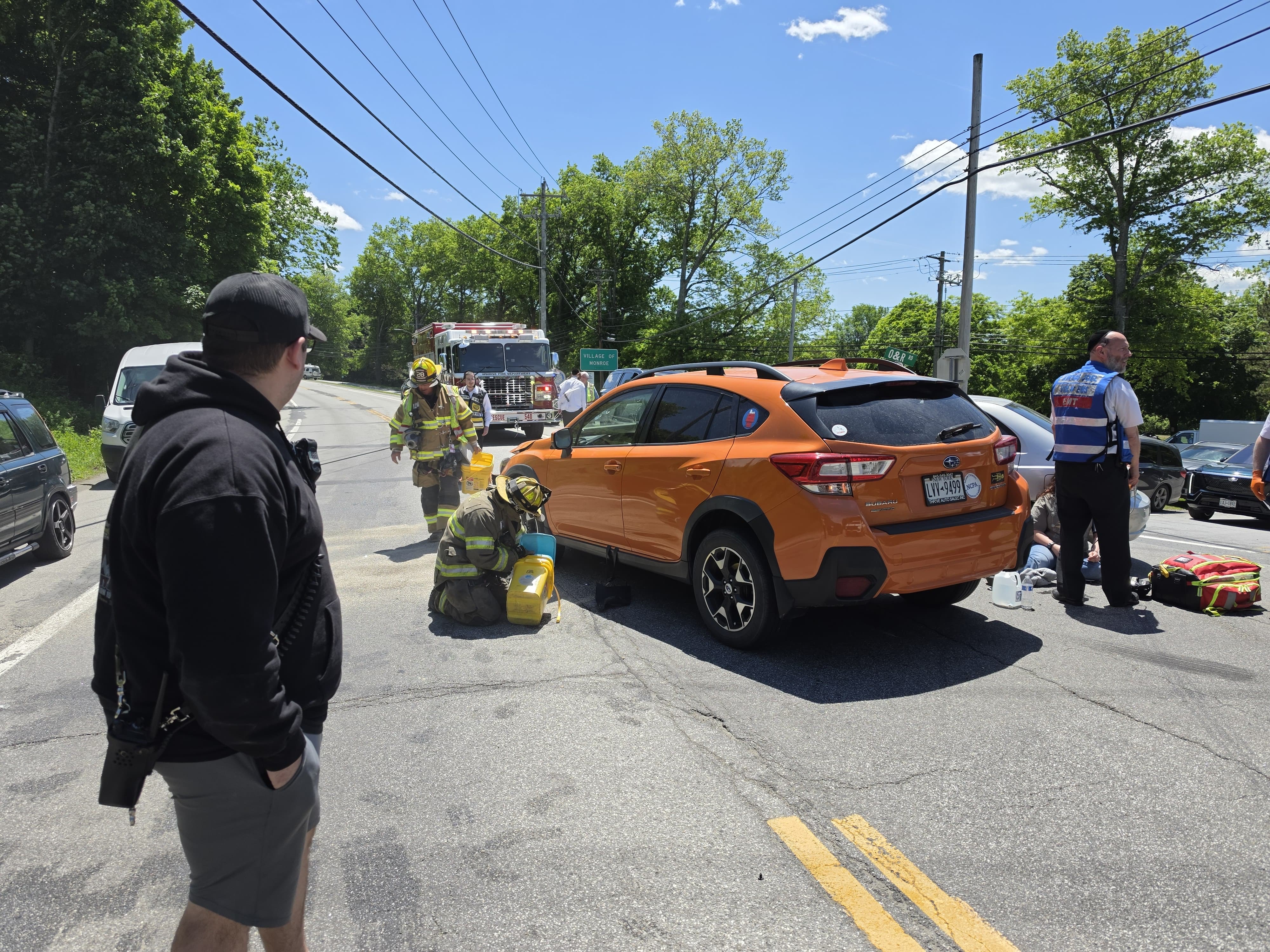 A Monroe firefighter laying down absorbent next to the orange Subaru Crosstrek while EMS tends to a patient and a fire engine stages up the road