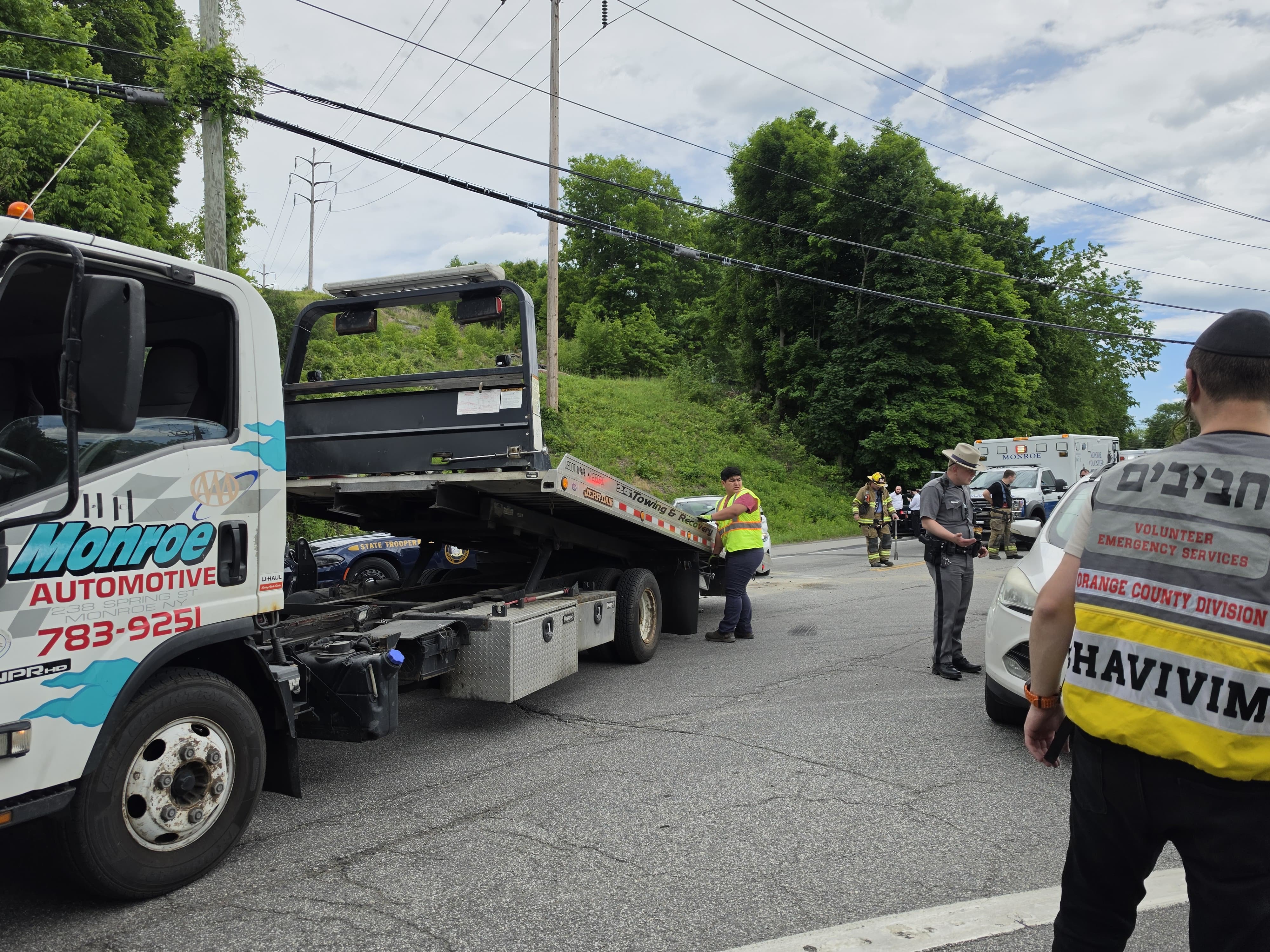 A Monroe Automotive flatbed tow truck on scene with firefighters, a state trooper, an ambulance, and a Chavivim member in an Orange County Division vest