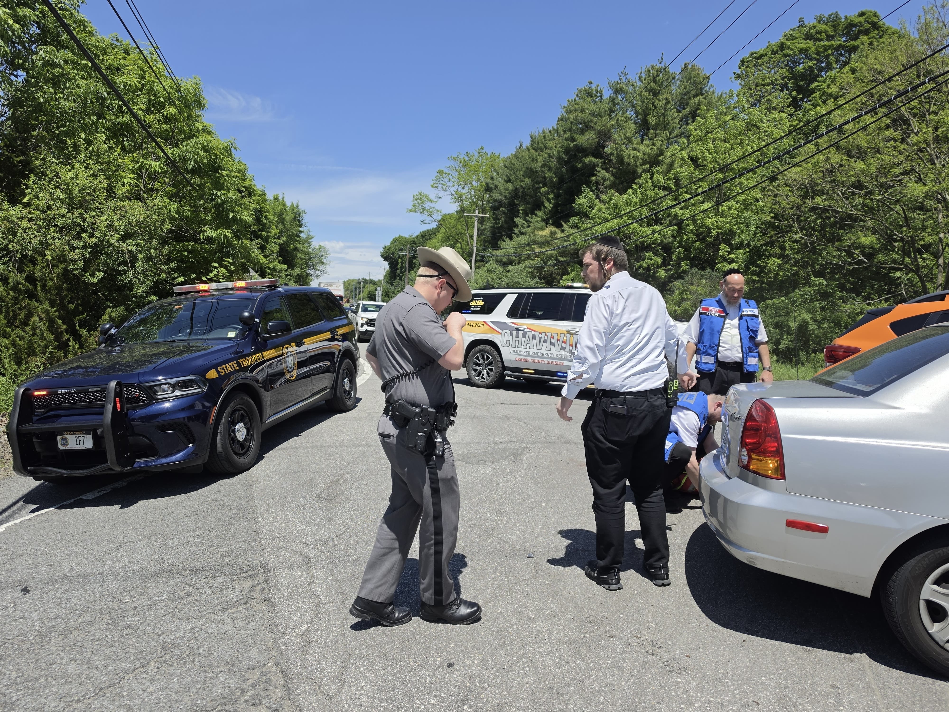 A New York State Trooper and a Chavivim member standing beside the damaged silver sedan, with a State Police SUV blocking the lane behind them